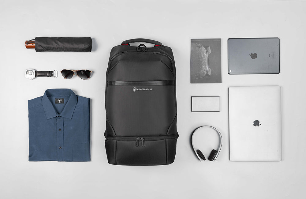 Black backpack with a shirt, umbrella, sunglasses, wallet, laptop, and headphones on a light gray background