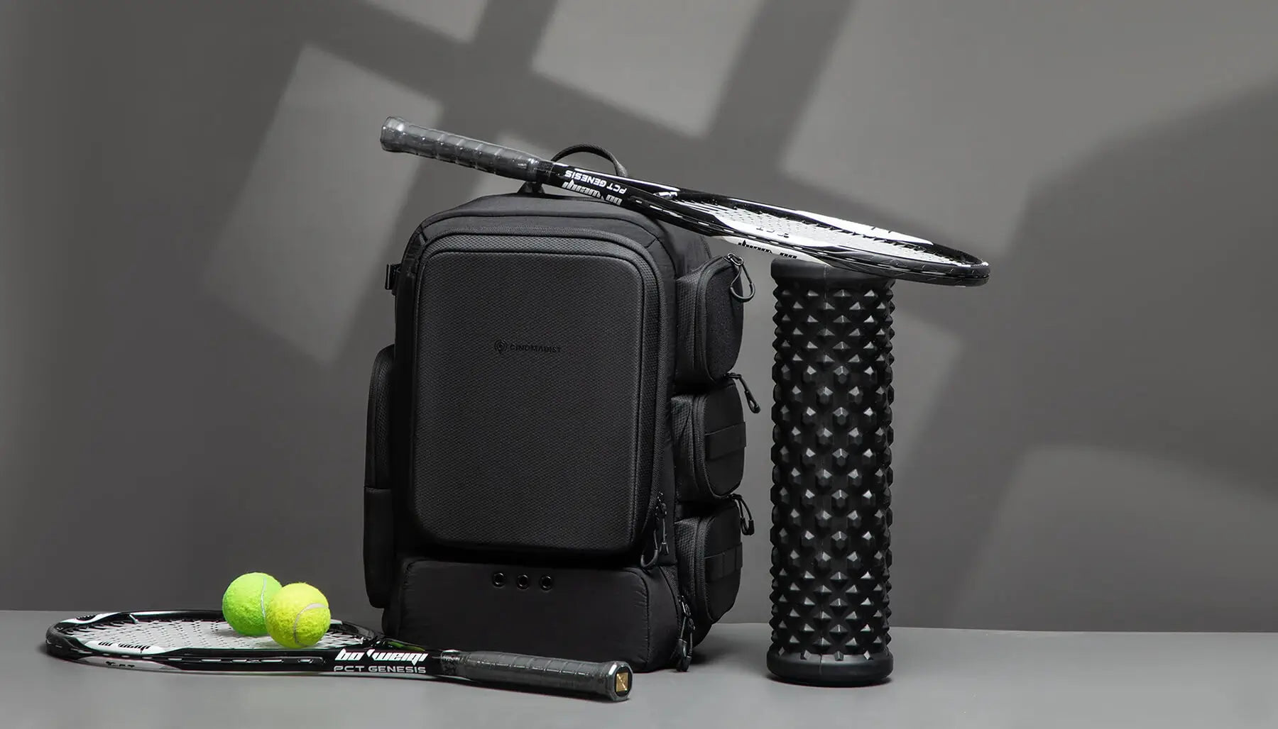 Black backpack with tennis racket, balls, and roller on a gray surface