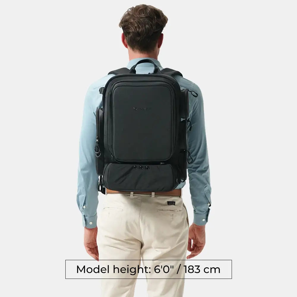 A man viewed from behind wearing a black backpack over a light blue dress shirt and cream pants, with a text overlay at the bottom stating 'Model height: 6'0" / 183 cm'.