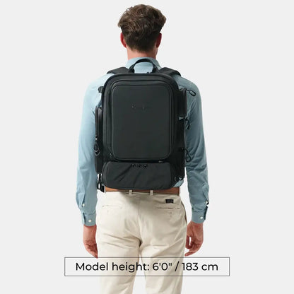 A man viewed from behind wearing a black backpack over a light blue dress shirt and cream pants, with a text overlay at the bottom stating 'Model height: 6'0" / 183 cm'.