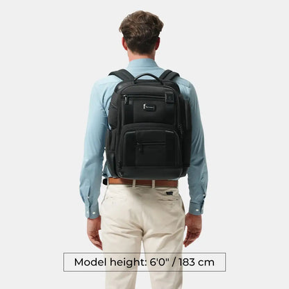A man viewed from behind wearing a black backpack, light blue dress shirt, and beige trousers against a light gray background. A text box at the bottom reads 'Model height: 6'0" / 183 cm'.