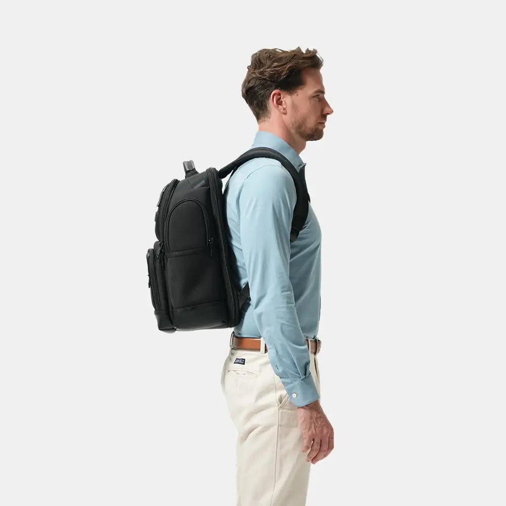 A man in a light blue dress shirt and cream trousers shown from the side, wearing a black backpack against a plain light gray studio background.